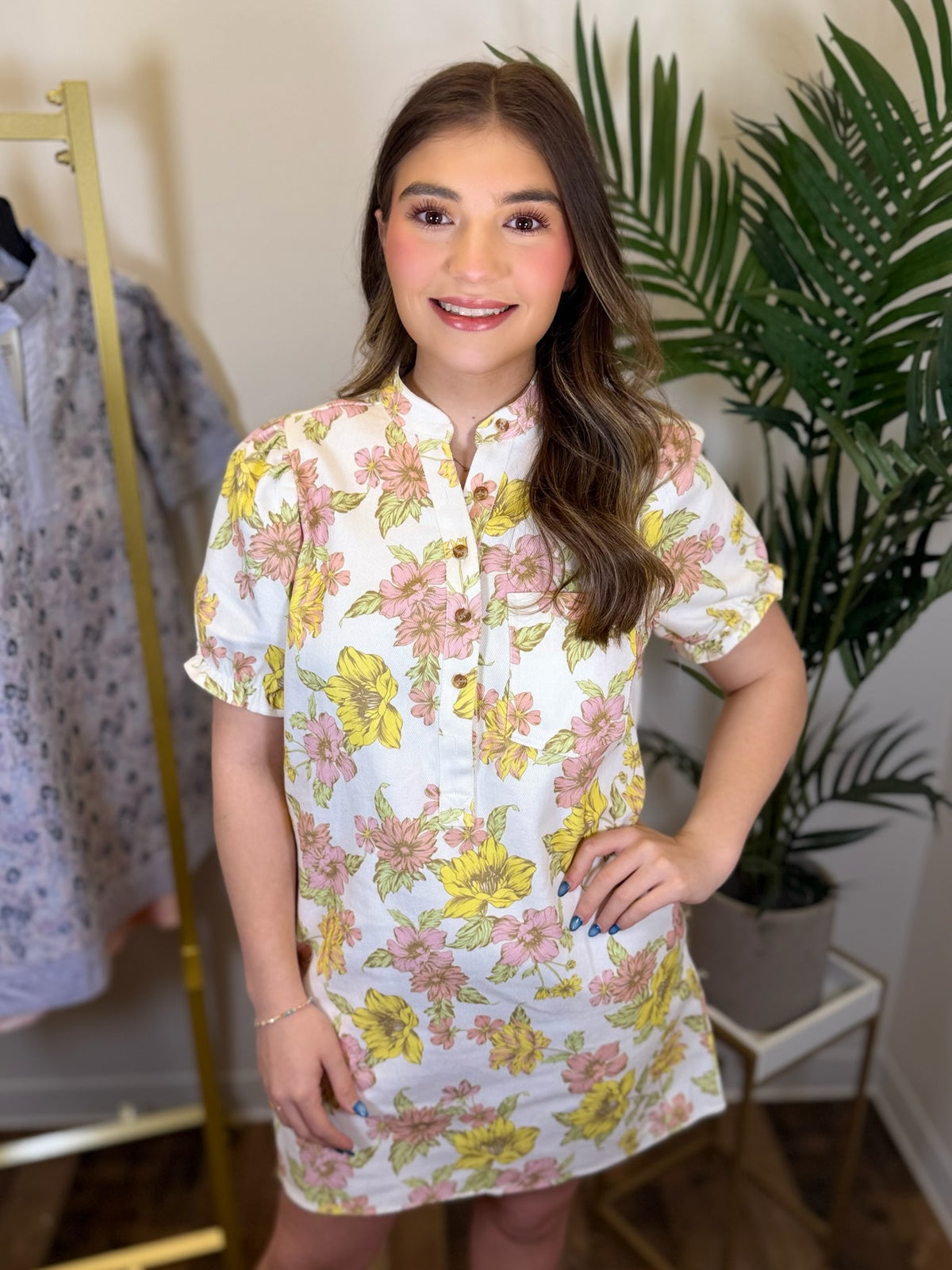Woman wearing a floral dress standing in a room with plants and a rack of clothes.