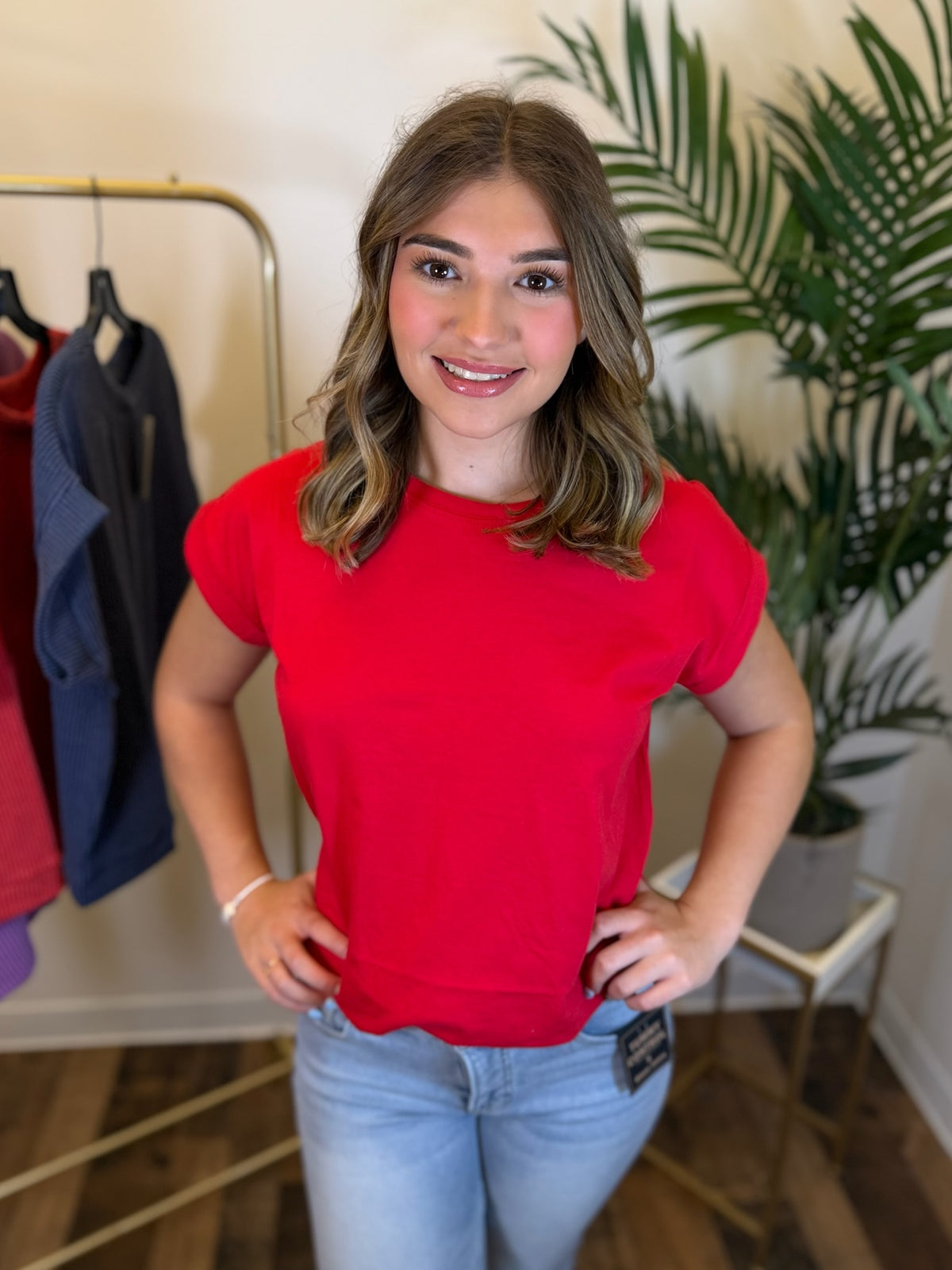 A woman wearing a bright red short-sleeve crewneck t-shirt and light blue jeans, standing in a boutique setting with a clothing rack and a green plant in the background.
