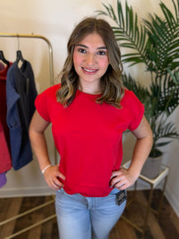 A woman wearing a bright red short-sleeve crewneck t-shirt and light blue jeans, standing in a boutique setting with a clothing rack and a green plant in the background.