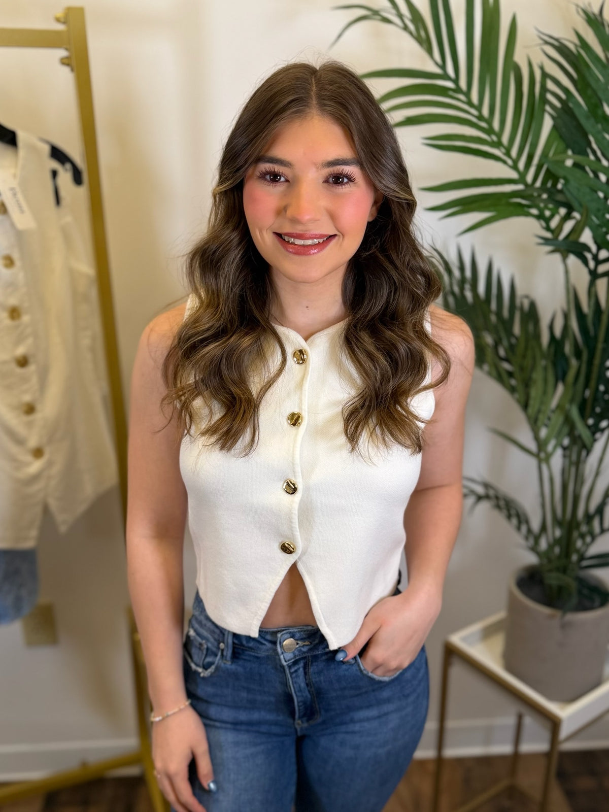 Woman wearing a white sleeveless top with gold buttons and blue jeans in a store setting.