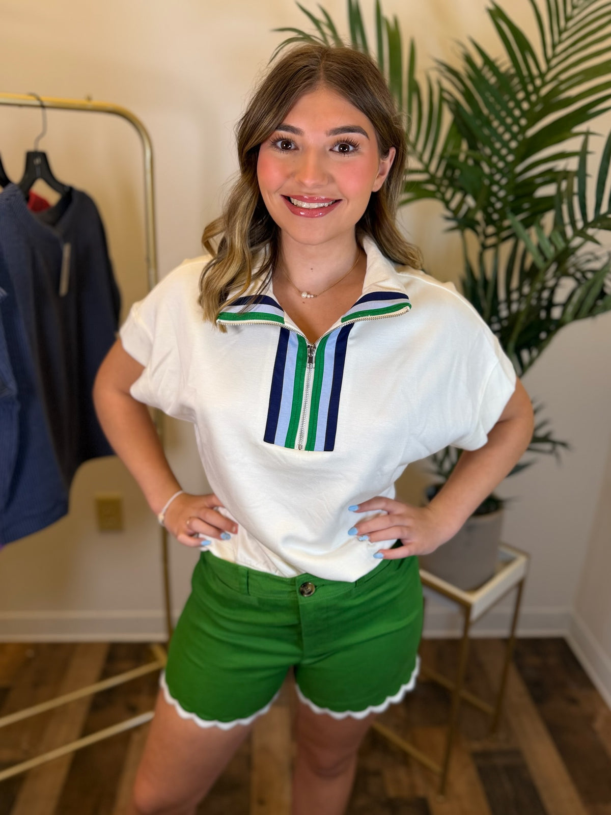 A woman modeling a white short-sleeve polo shirt with green and navy striped collar details, paired with bright green shorts, standing in an indoor boutique setting.