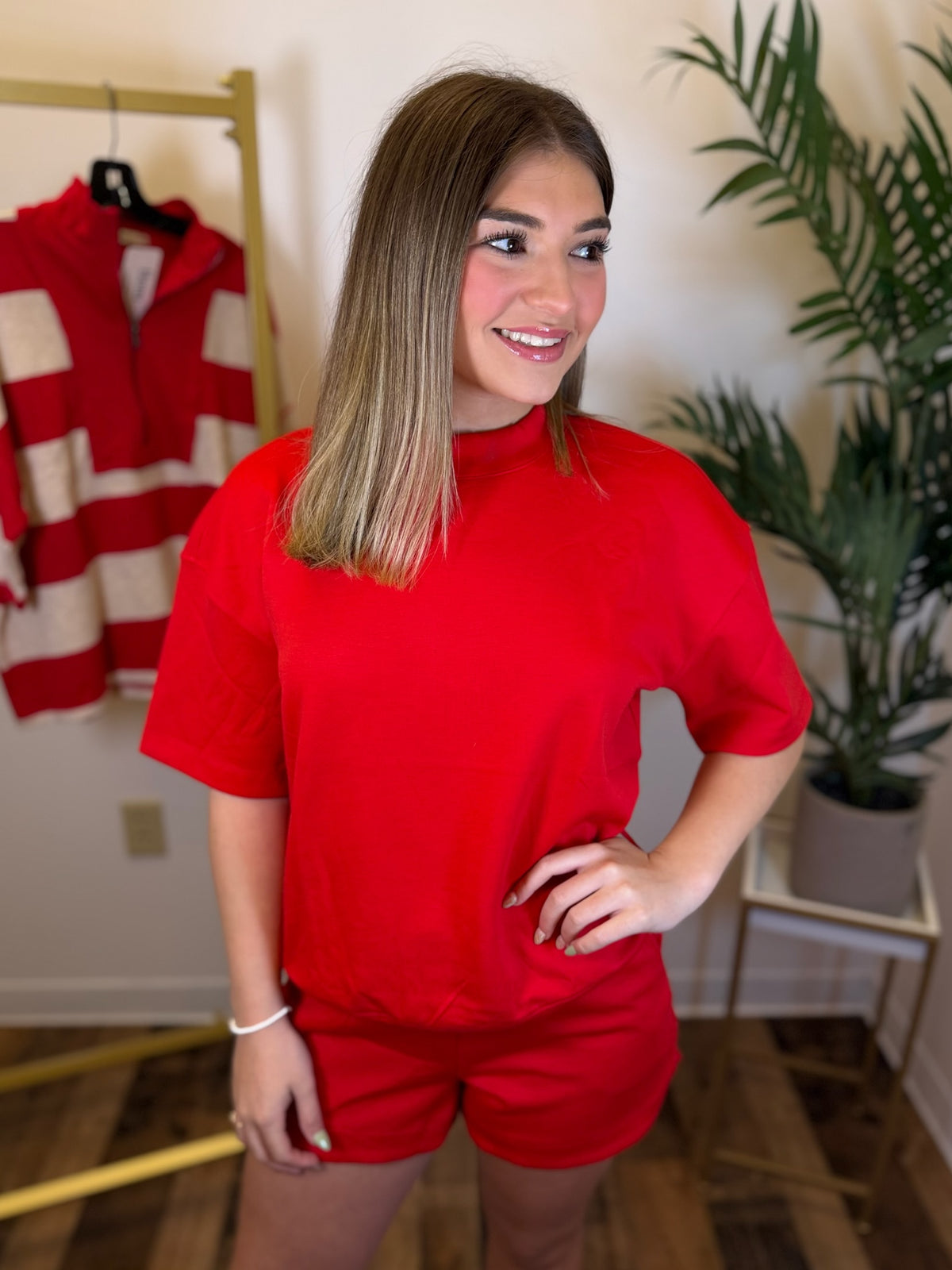 A woman modeling a bright red short-sleeve crewneck t-shirt and matching red shorts in an indoor setting with a clothing rack and a potted plant in the background.