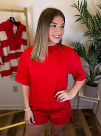A woman modeling a bright red short-sleeve crewneck t-shirt and matching red shorts in an indoor setting with a clothing rack and a potted plant in the background.