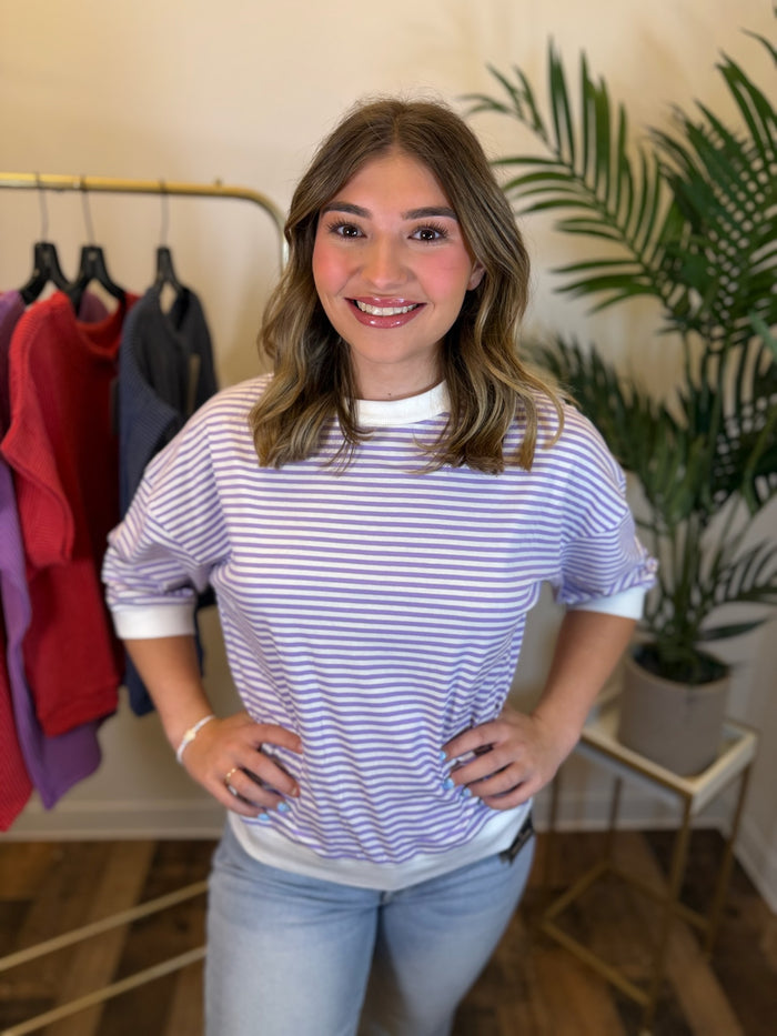 A woman smiling and posing in a light purple and white horizontally striped t-shirt with white contrast trim sleeves and hem, paired with light blue jeans, in a boutique setting with a clothing rack and a potted plant in the background.