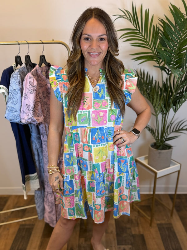 Woman wearing a short colorful dress with ruffled cap sleeves and split neckline standing in front of a clothing rack with plants in the background