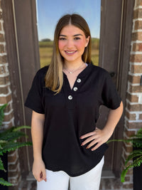 A woman wearing a black short-sleeve ribbed henley top with three button plackets, paired with white pants, standing in front of a brick door.