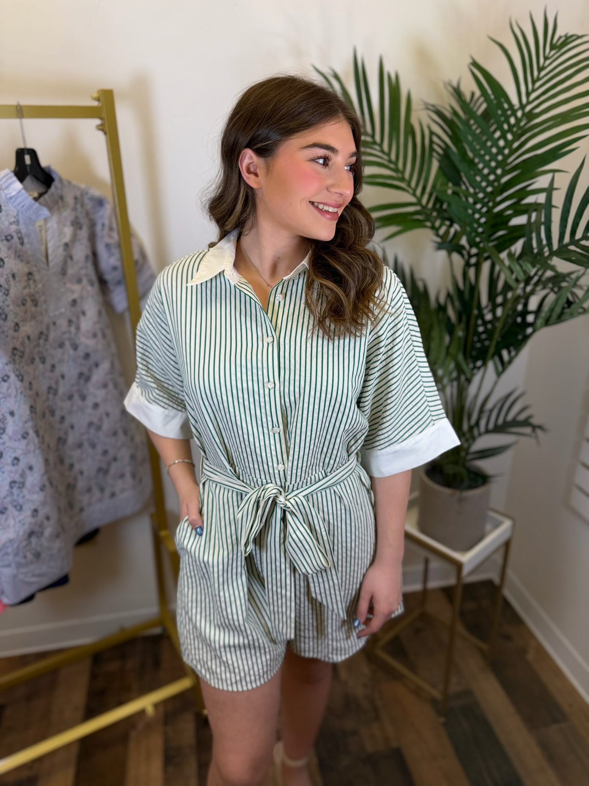 Woman wearing a short sleeve green and white striped romper with tie waist in a room with plants and clothing racks.