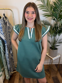 Woman wearing an olive v neck dress with ruffled cap sleeves and contrast trim details standing in a room with a clothing rack in the background.