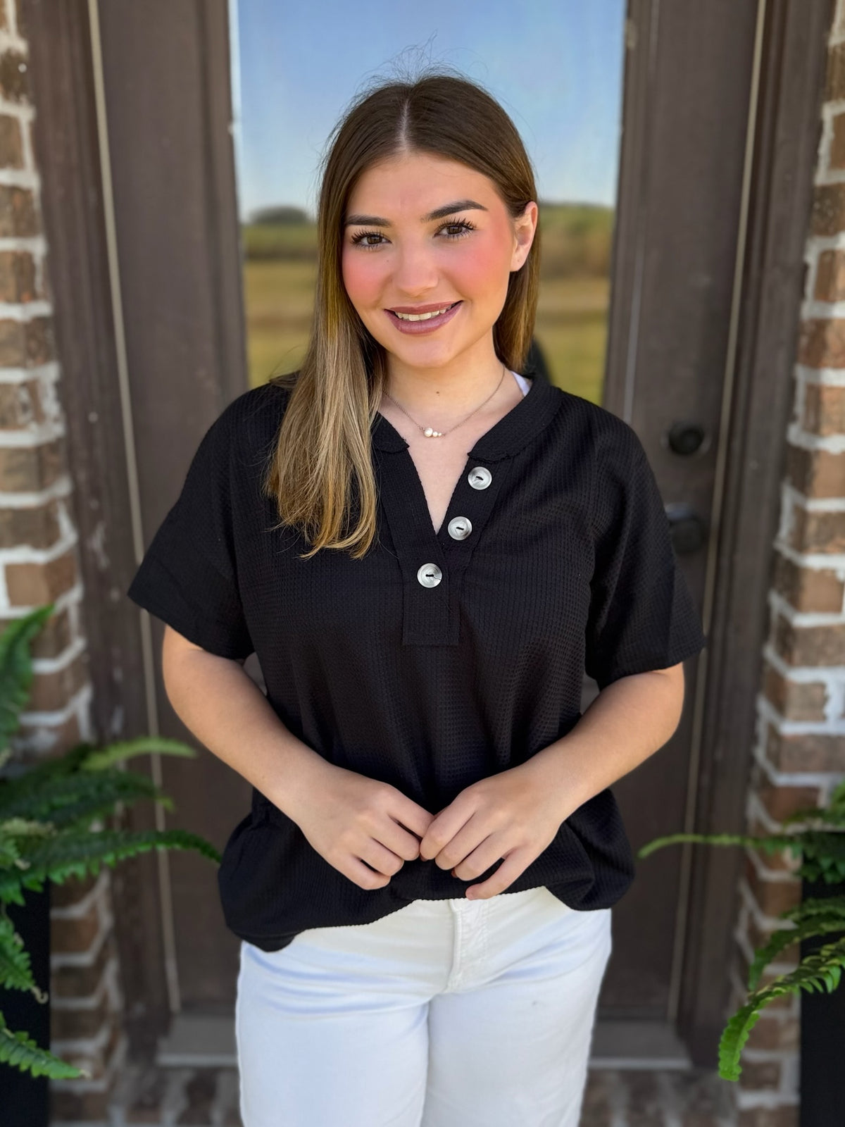 A woman wearing a black short-sleeve top with a three-button placket and white pants, standing in front of a brick doorway with plants visible on both sides.