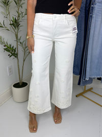Person wearing cream wide leg jeans with 3d floral embellishments in a room with a plant and clothing rack.
