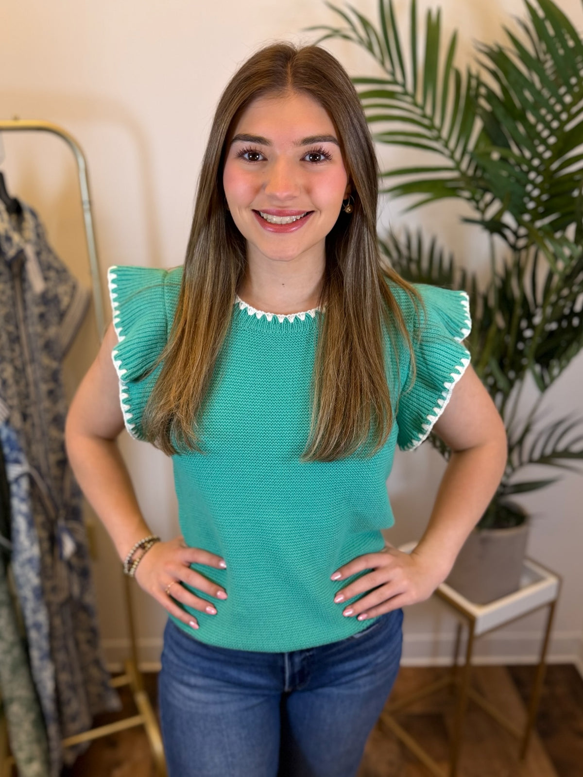 Woman wearing a teal top with ruffled sleeves in a room with plants and a mirror.
