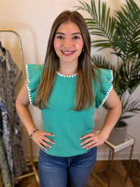 Woman wearing a teal top with ruffled sleeves in a room with plants and a mirror.