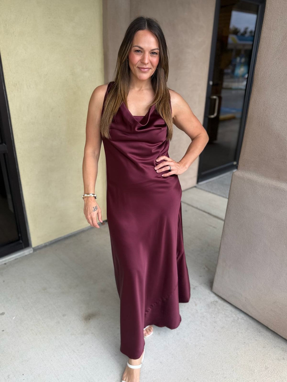 Burgundy satin midi dress with spaghetti straps and draped neckline worn by a woman standing in front of beige building.