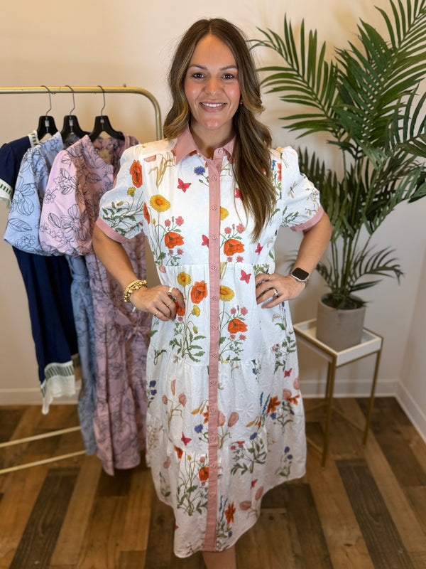 Woman wearing a white floral dress with short sleeves and a tiered midi fit standing in a clothing store with racks of dresses and plants in the background.