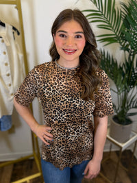 Person wearing a mesh leopard print top with short sleeves and ruffled trim in a room with plants and clothing racks.