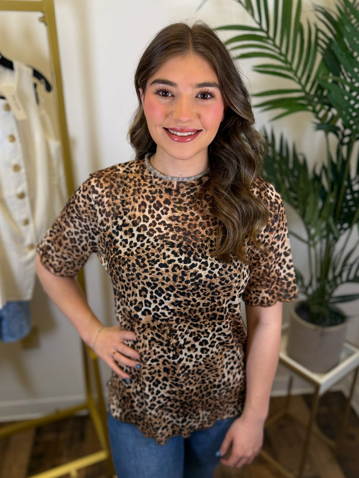 Person wearing a mesh leopard print top with short sleeves and ruffled trim in a room with plants and clothing racks.