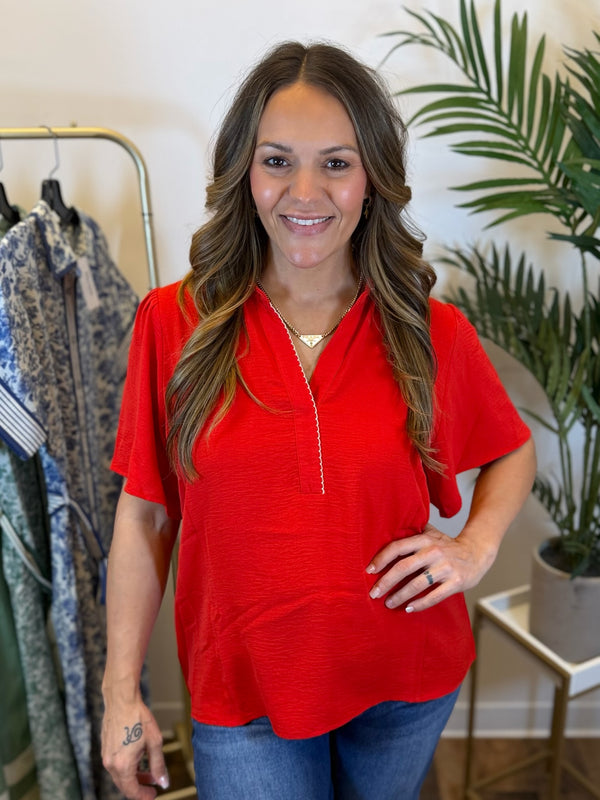 Woman wearing a red blouse with short sleeves and collared v neck with scalloped trim in a clothing store setting