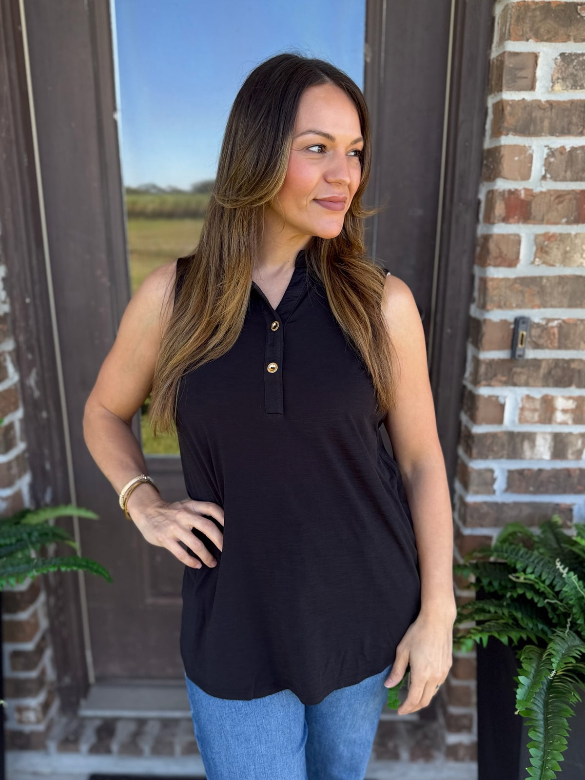 A woman wearing a sleeveless black polo-style top with gold buttons, paired with blue jeans, standing in front of a brick door.