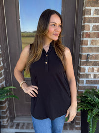A woman wearing a sleeveless black polo-style top with gold buttons, paired with blue jeans, standing in front of a brick door.