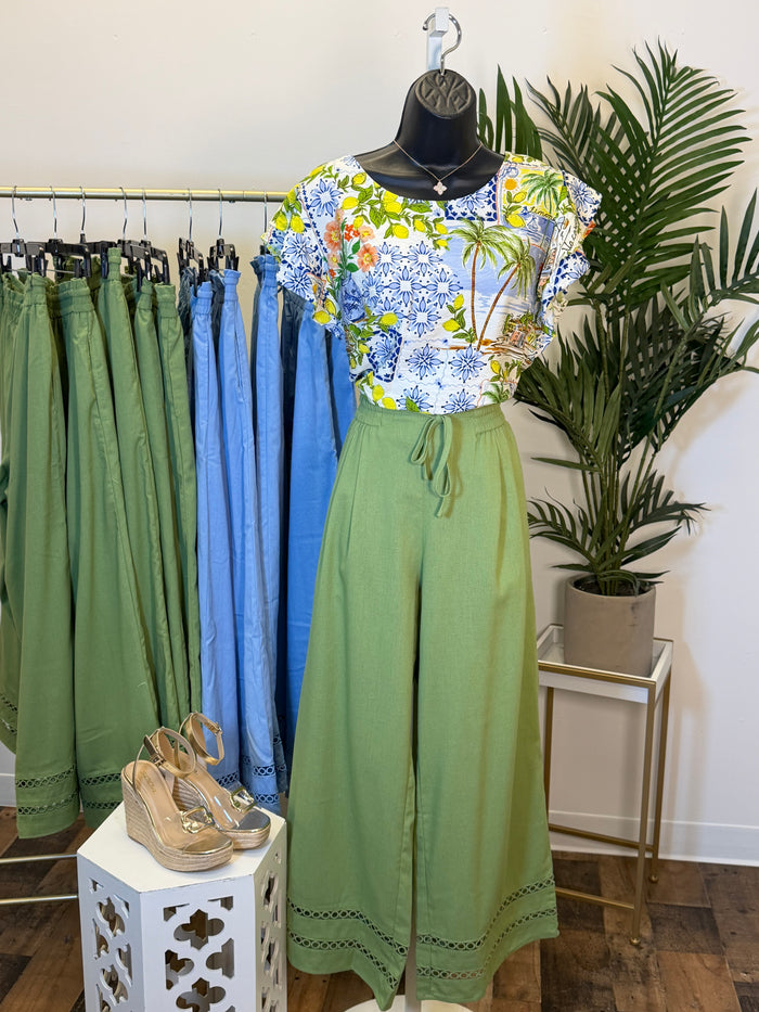 Mannequin wearing white cap sleeve blouse with tropical print and green bottoms with clothing rack, shoes, and plant in the background.