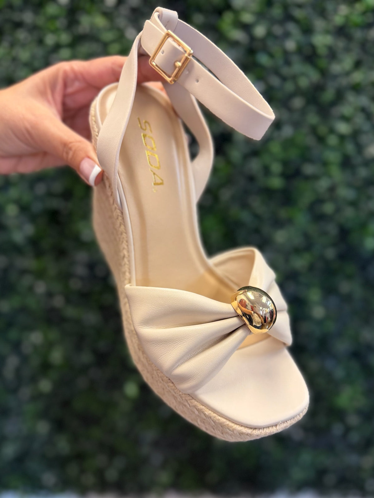 Beige sandal with a decorative bow held against a green leafy background