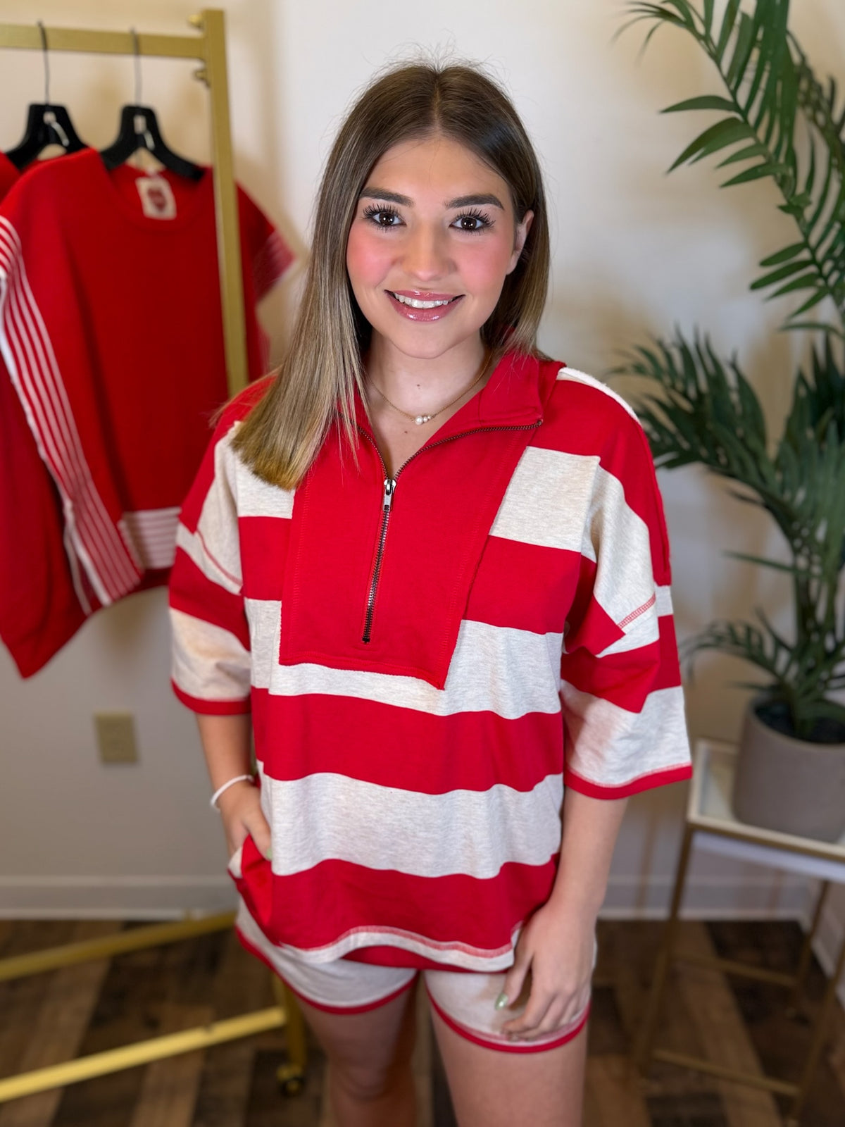 A woman modeling a red and white striped short-sleeve half-zip pullover with matching striped shorts in an indoor boutique setting.