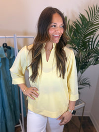 Woman wearing a yellow blouse with ruffled neckline standing in a room with a clothing rack and plant.