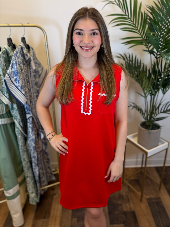Woman in a red sleeveless dress with white ric rac trim and zip neckline standing in a room with clothing and plants.