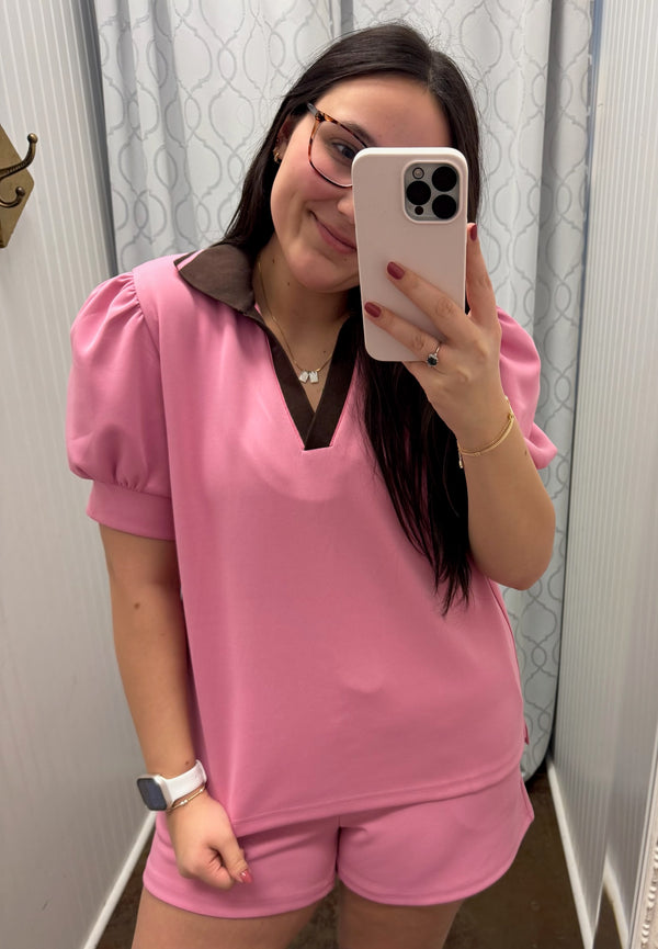 Woman taking a mirror selfie wearing a pink outfit with puff sleeves and brown trim.