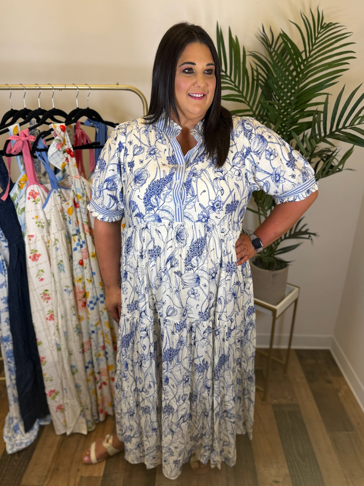Woman wearing a blue and white floral dress standing in a room with clothing racks and plants.