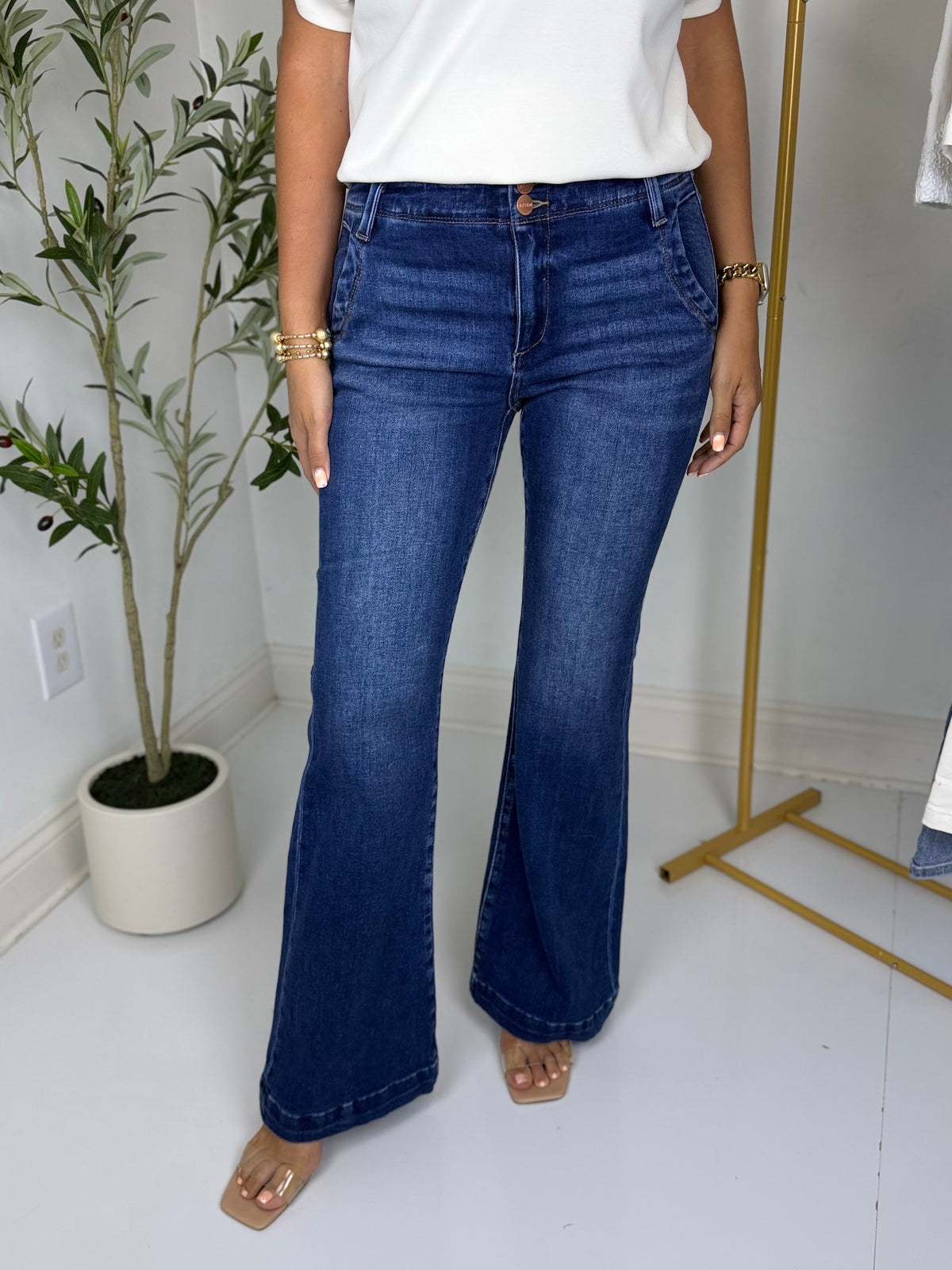 Person wearing dark blue flared jeans with trouser style pockets and a white top in a room with a plant and coat rack.