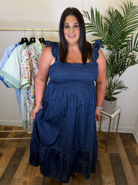 Woman wearing a denim blue maxi dress in a room with plants and clothing racks.