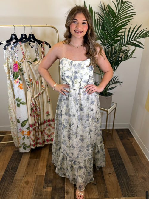 Woman wearing a strapless floral dress with corset inspired bodice and midi length standing in a room with a rack of dresses and a plant.