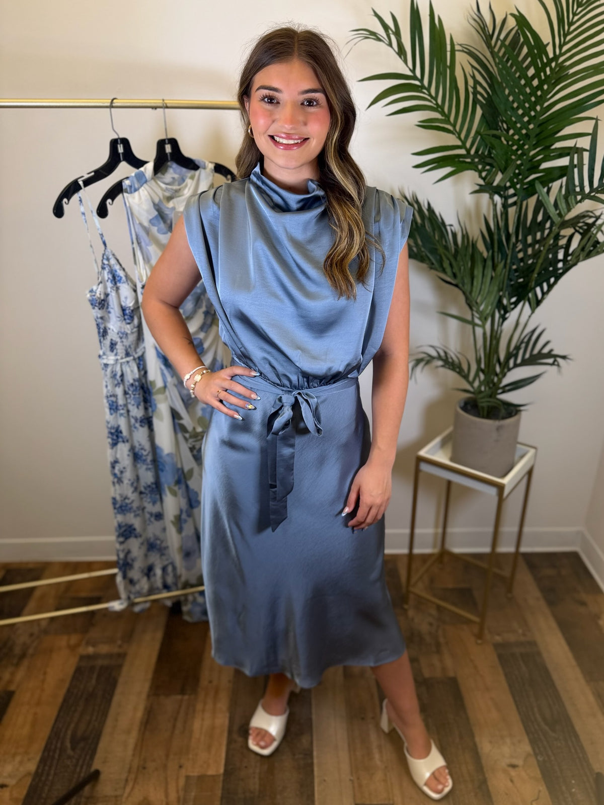 Woman wearing a blue midi dress standing in a room with a plant and clothing rack.