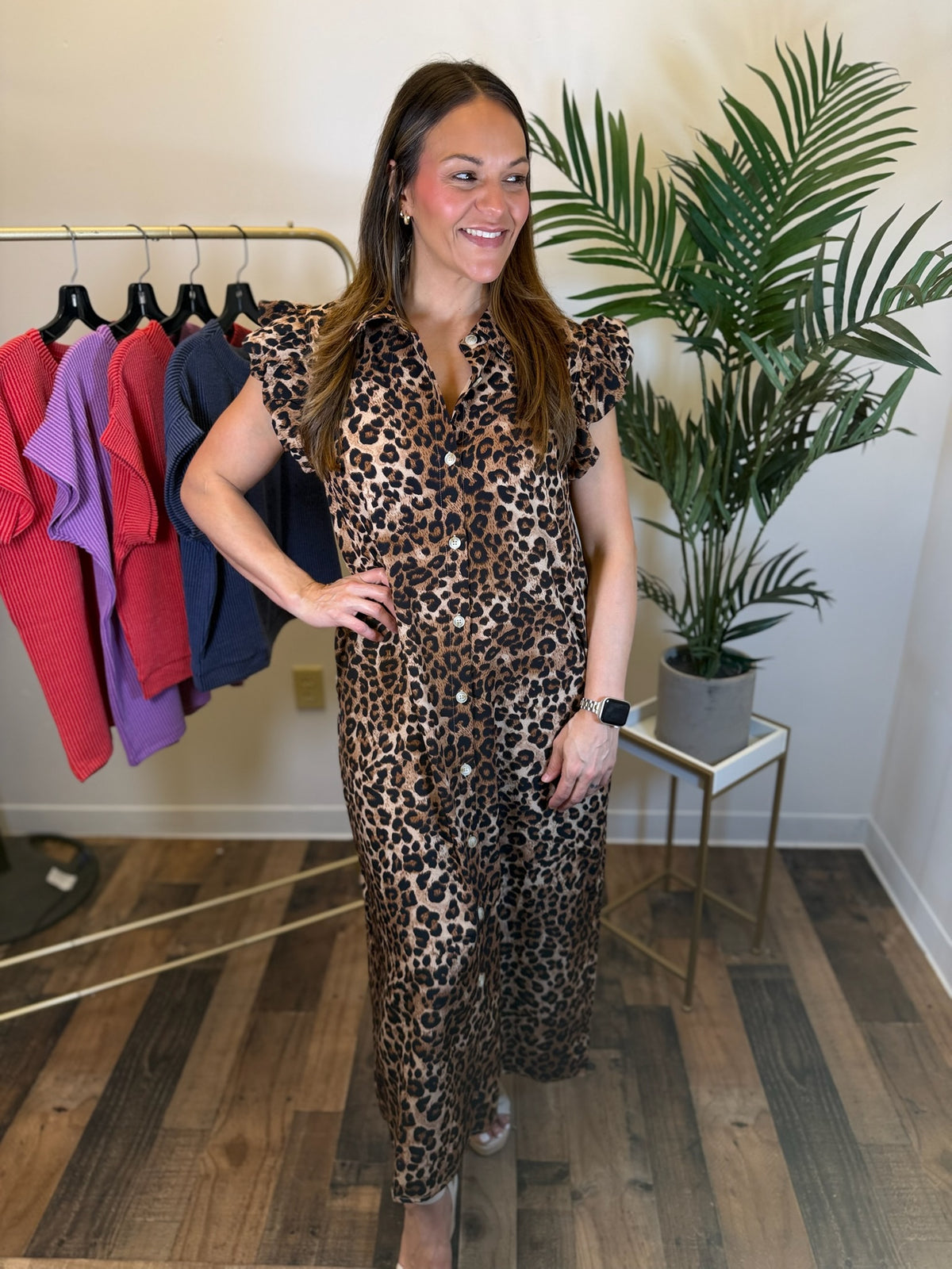 A woman modeling a leopard print maxi dress in a boutique setting, standing next to a clothing rack and a potted plant.