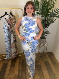 Woman wearing a white and blue floral dress with ruching standing in a room with clothing racks and plants.