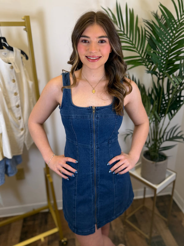 Woman wearing a blue denim dress in a room with plants and clothing racks.