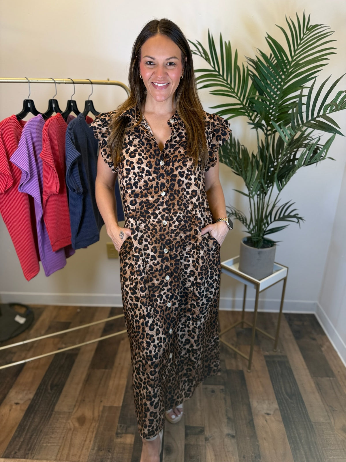 A woman modeling a leopard-print maxi dress with short flutter sleeves and side pockets, standing in a boutique setting with a clothing rack and a potted plant in the background.