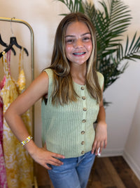 Young woman wearing a light green sleeveless button-up top and blue jeans, standing in a room with a rack of clothes and a plant.