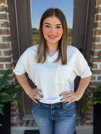 A young woman with long brown hair stands in front of a brick doorway wearing a white short-sleeve waffle-knit top with a chest pocket and blue jeans.