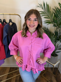 A woman wearing a pink vertically striped button-up blouse with puff sleeves and a ruffled hem, standing in a boutique setting with a clothing rack and potted plant in the background.