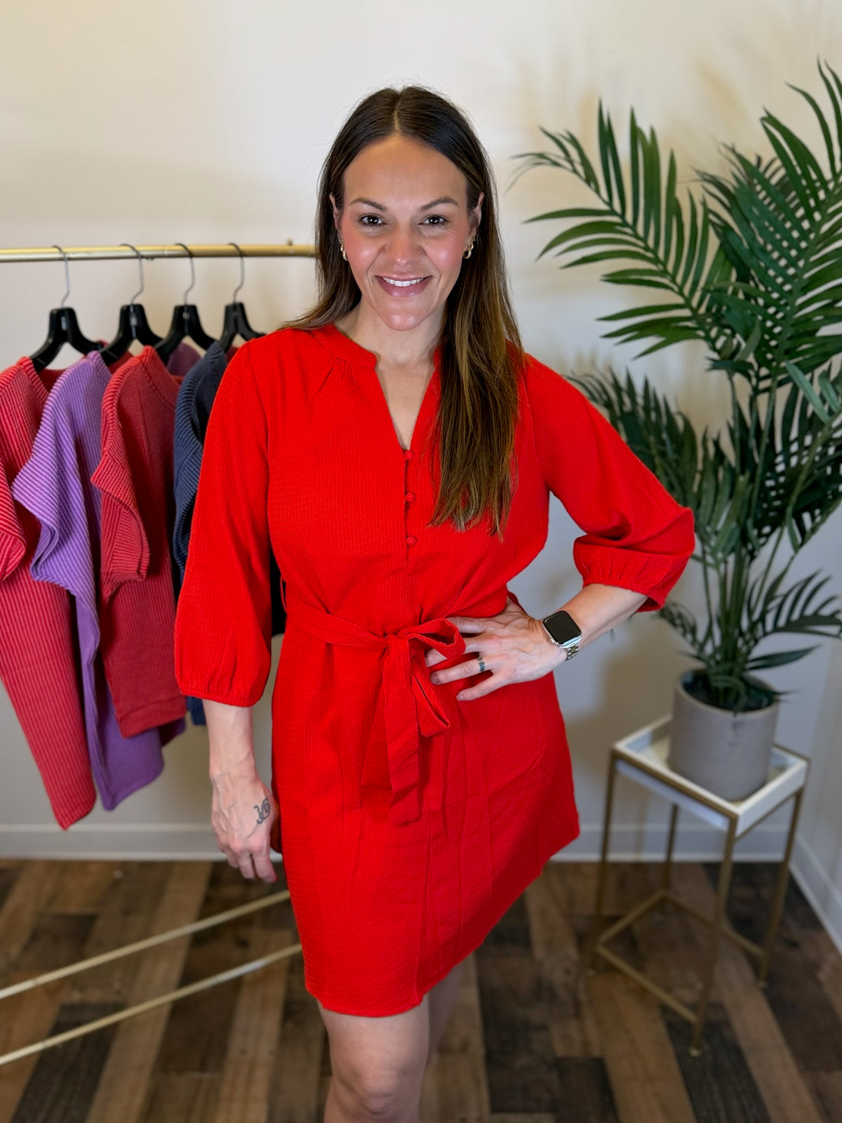 A woman modeling a bright red mini dress with puff sleeves and a self-tie waist belt, standing in a boutique setting with a clothing rack and a potted plant in the background.