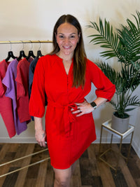 A woman modeling a bright red mini dress with puff sleeves and a self-tie waist belt, standing in a boutique setting with a clothing rack and a potted plant in the background.