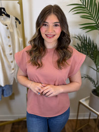 Woman wearing a pink top with round neck and blue jeans standing in a room with a plant and clothing rack.