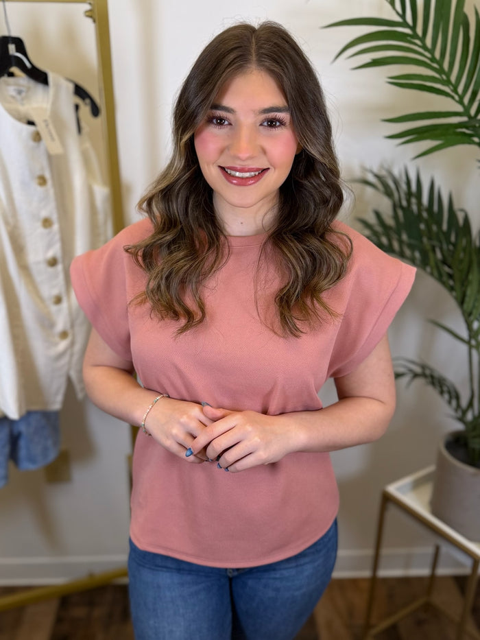 Woman wearing a pink top with round neck and blue jeans standing in a room with a plant and clothing rack.