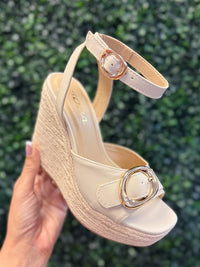 Beige wedge sandal with a decorative buckle held against a green leafy background