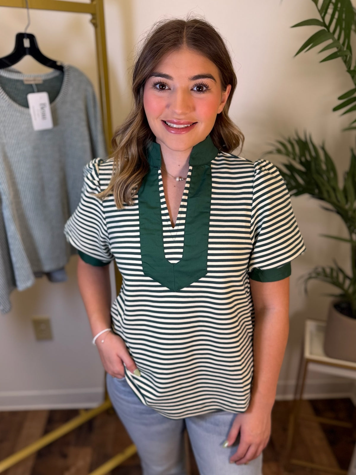 A woman modeling a short-sleeve top with green and white horizontal stripes and a solid green V-neck trim, paired with light blue jeans in an indoor boutique setting.