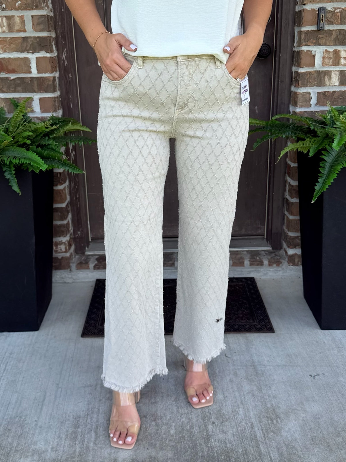 A person wearing light beige textured straight leg jeans with a subtle geometric pattern and frayed hems, paired with a white top and clear heeled sandals, standing on a concrete porch in front of a brick door.