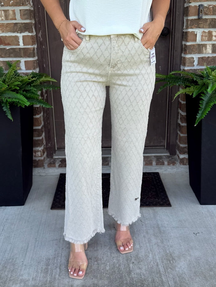 A person wearing light beige textured straight leg jeans with a subtle geometric pattern and frayed hems, paired with a white top and clear heeled sandals, standing on a concrete porch in front of a brick door.