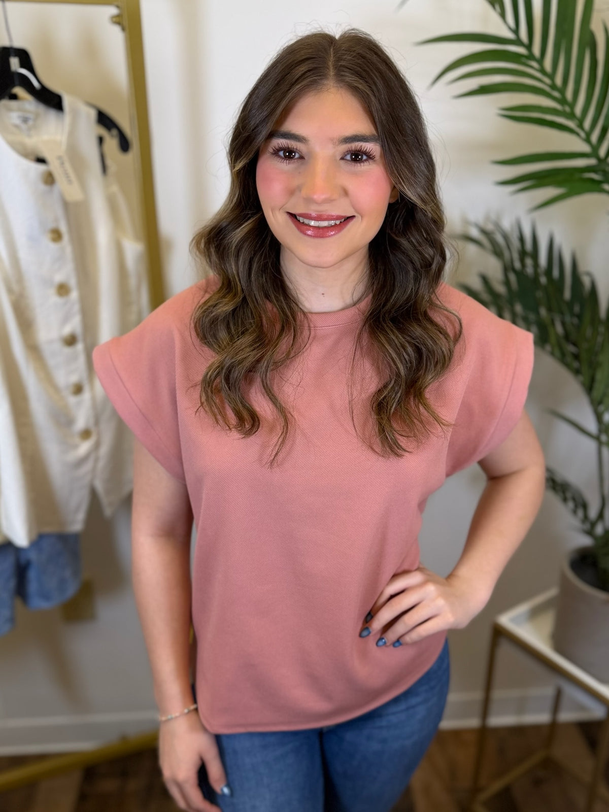 Woman wearing a casual pink top and blue jeans standing in a store setting with clothing and plants in the background.